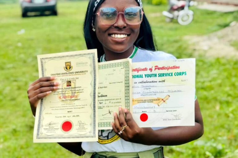 Smiling young woman in glasses and NYSC uniform holding up her university degree certificate, NYSC certificate of national service and an NYSC certificate of participation outdoors